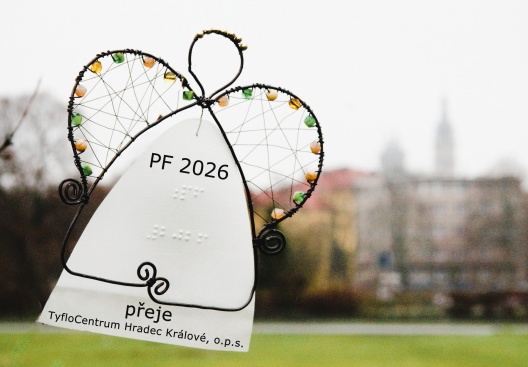 Drátěný andělíček má bílou košilku, na které je napsáno černotiskem i Braillovým písmem: PF 2026, přeje TyfloCentrum Hradec Králové, o.p.s. Andělíček se vznáší v prostoru, v pozadí je zamlžený, neostrý pohled na panorama historického centra Hradce Králové.